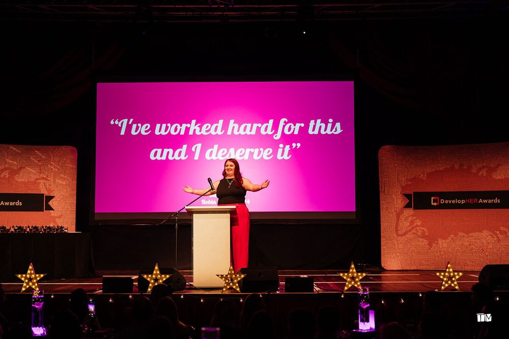 A woman stands on a stage in front of a projector screen with her arms open wide, looking at the audience. Behind her the projector screen displays a quote in white text on a bright pink background: “I’ve worked hard for this and I deserve it”. It is a quote from the Barbie movie.