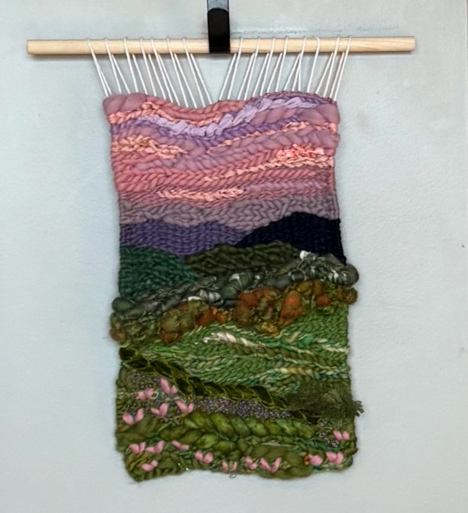 A woven wall hanging on a wooden dowel showing green rolling hills with dark purple mountains behind and a lavender and peach sunset sky