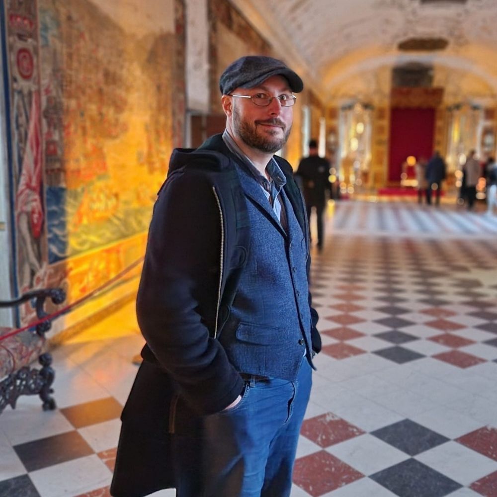 A picture of our group member Jordan Gault, taken in the caste in Copenhagen. You can see a bearded man with glasses standing in a grand, ornately decorated hallway with a checkered marble floor. He has dark brown hair and is smiling. He is wearing a flat cap, blue vest over a shirt, dark trousers, and a dark jacket with one hand in his pocket. Behind him, the hallway is lined with large, colorful tapestries and elegant furniture, leading to a distant area with a red throne-like seat. The lighting is warm and soft, giving the scene a historic and majestic ambiance. And yes, he looks like a mixture of an oldtimey academic and a wizard (in a good way).
