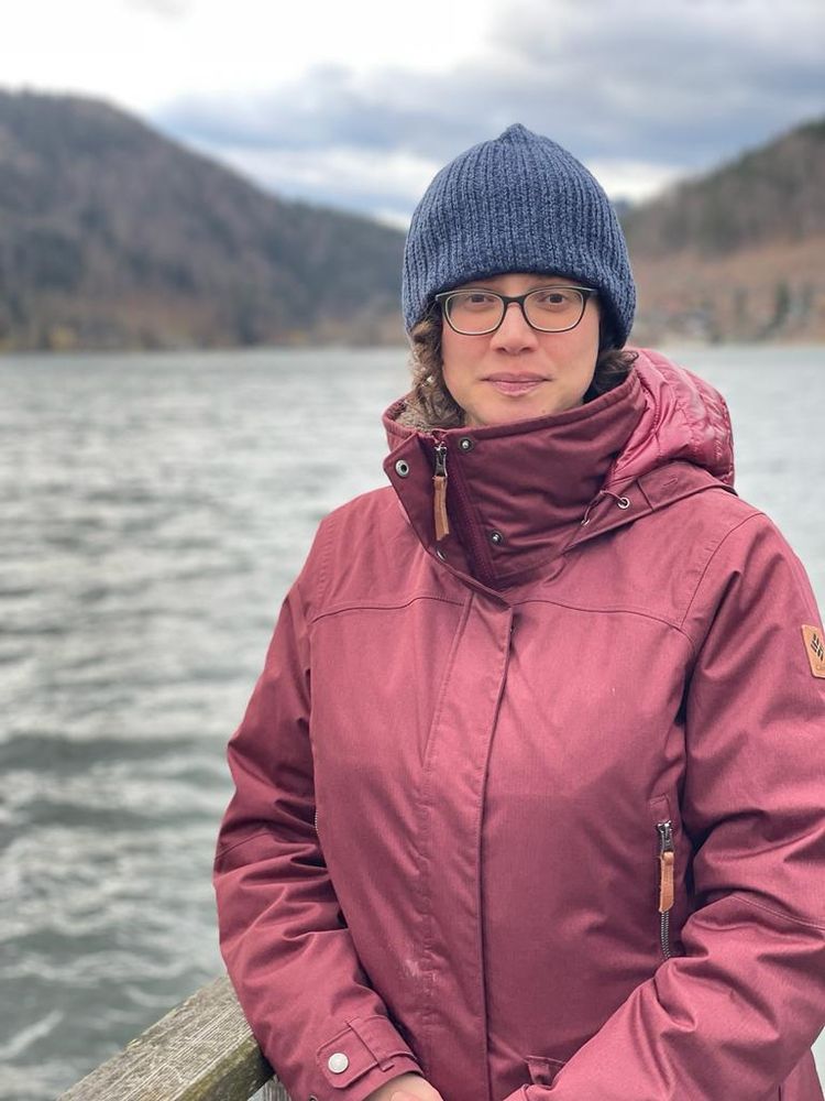 A picture of our group member Laura Barth. She is wearing a maroon winter jacket, navy blue knitted beanie, and glasses. You can see her dark curly hair sticking out under the beanie. Laura is standing outdoors on a wooden pier by a calm body of water. She has a slight, friendly smile and is looking at the camera. Behind her are forested hills and a cloudy sky, creating a serene atmosphere.