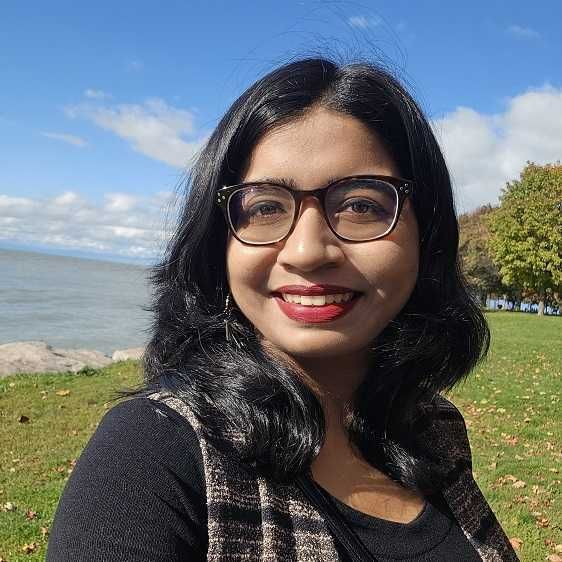 A picture of our group member Srilena Kundu. The picture shows a woman with shoulder-length black hair, wearing glasses, a black top, and a patterned vest. She smiles brightly at the camera. She is outdoors on a sunny day, standing on a grassy area with trees and a large body of water visible in the background. The sky is clear with scattered white clouds.