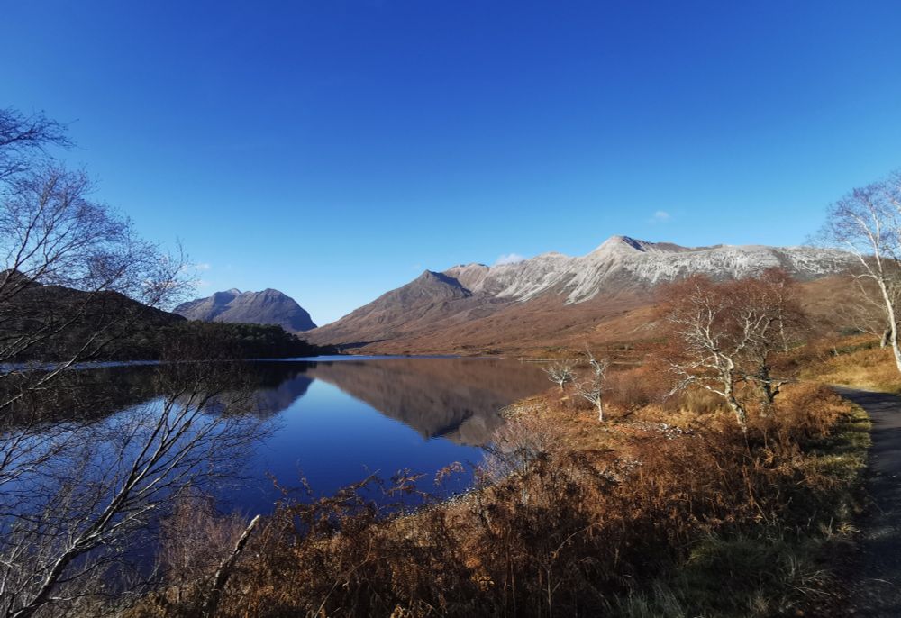 Reflection of distant mountains & blue sky with bare trees along the shoreline. 