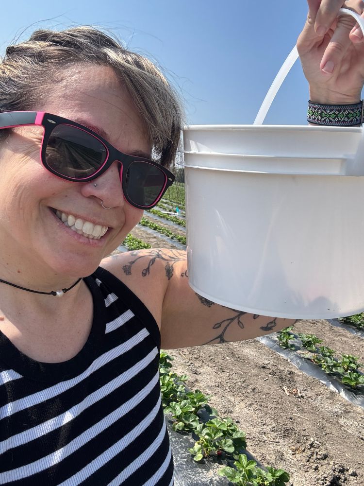 Happy Rebekah, wearing sunglasses and a black striped tank, holding a bucket of berries