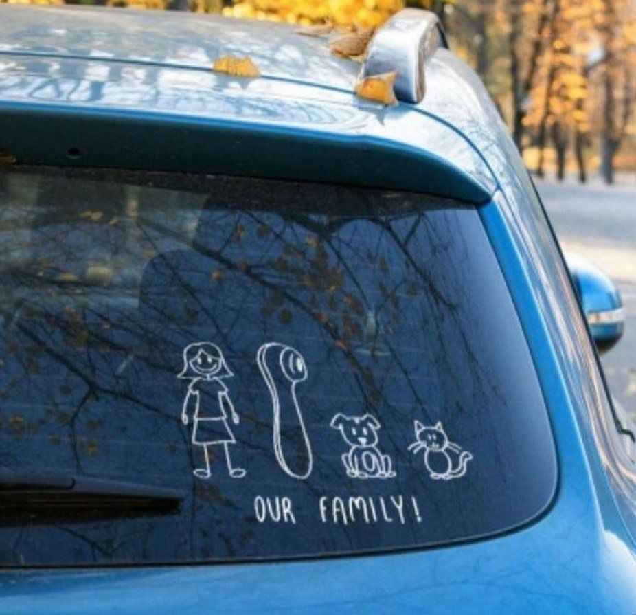 A white vinyl car decal on a dark window depicts a simple stick-figure woman standing upright with a smiling face. To her immediate right is a vibrator. Next to that, there is a small stick figure dog with pointed ears and a wagging tail, followed by a stick figure cat sitting with an arched tail. The decal mimics traditional stick family stickers but humorously replaces a spouse & kids with pets and a sex toy.