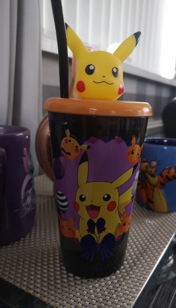 A black plastic cup with a design of Pikachu wearing a little black bowtie and surrounded by little orange pumpkin Pikachu and balloons. The lid is orange with a black straw, and a 3D Pikachu head on top