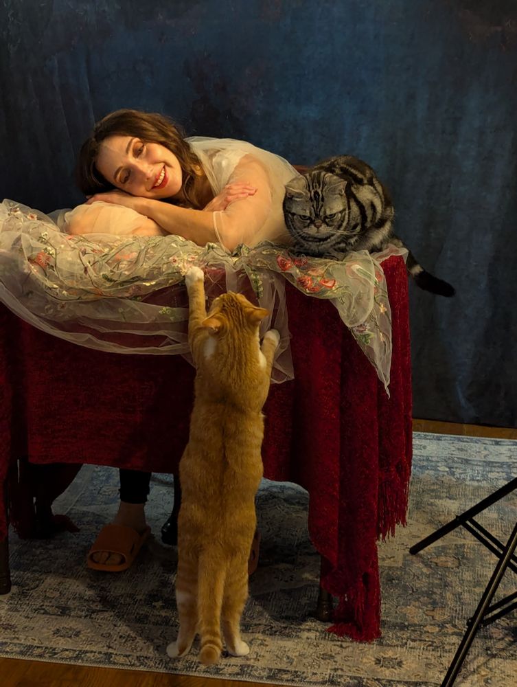 Two cats - one on the table, one trying to climb on the table - posing with a model 