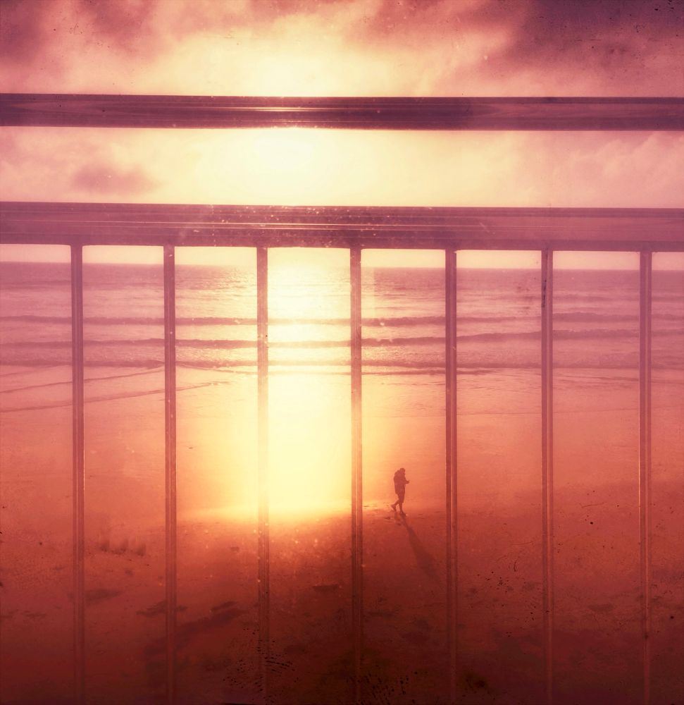 A warm, dreamy photo of a sunset over the ocean, viewed through vertical metal bars of a railing. The sun glows intensely, casting golden light on the beach below, where a lone person stands near the water’s edge, their long shadow stretching across the sand. The image has a nostalgic, vintage feel with soft pink and orange tones.
