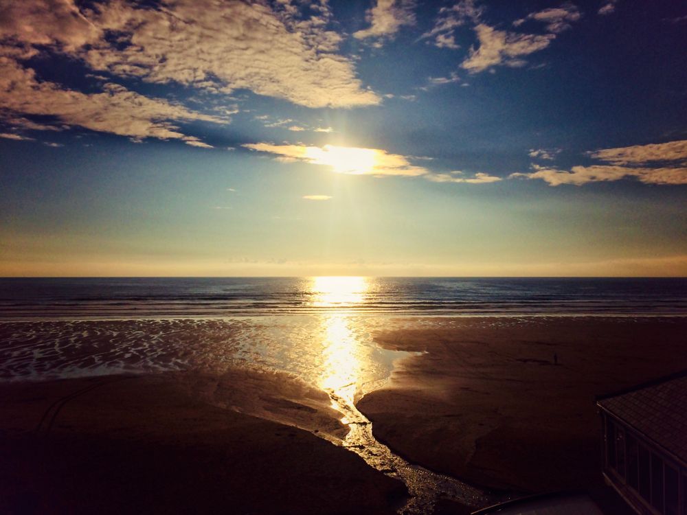A serene coastal scene during sunset with the sun low in the sky, casting a golden reflection across the ocean and wet sand. The tide is out, revealing patterns in the sand and a small stream leading toward the sea. Soft clouds scatter across the darkening blue sky. 