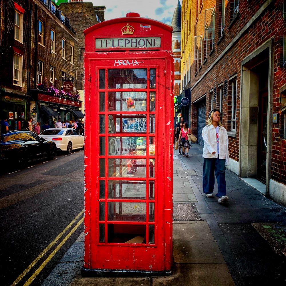 A classic red British telephone box stands prominently in the middle of a lively London street. The booth shows signs of wear and graffiti. Surrounding it are pedestrians, including a young woman in casual streetwear, wearing headphones walking past on the pavement. The buildings lining the street are made of traditional red brick, and cars line the narrow road, blending historic charm with contemporary life.
