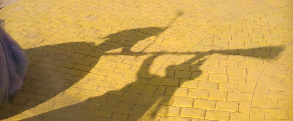 Image de "Wicked for good" : les ombres d'Elphaba et Glinda se battant se reflètent sur la route en briques jaunes 