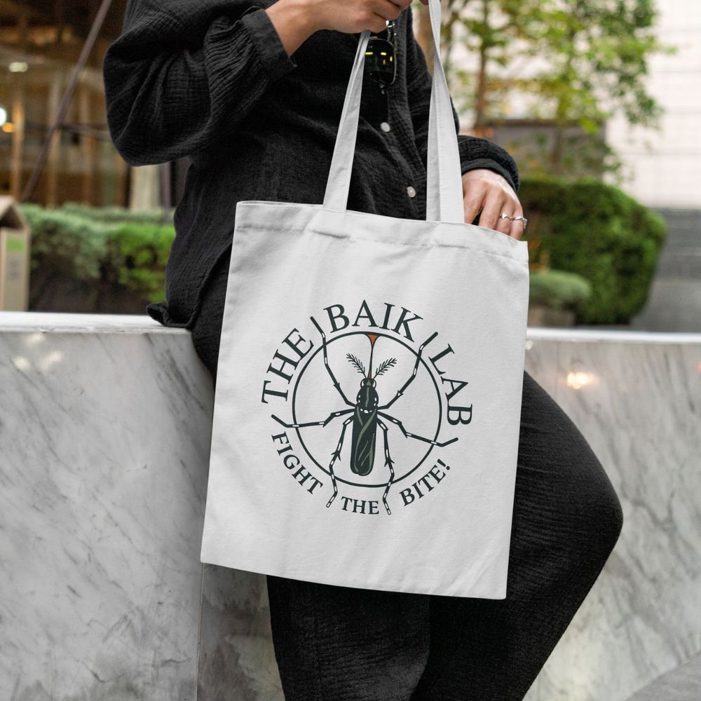 A person dressed in all black, leaning against a marble wall. The person is holding a cream colored tote bag with a circular mosquito logo that reads "The Baik Lab, Fight the Bite." In the background are green bushes and trees. 