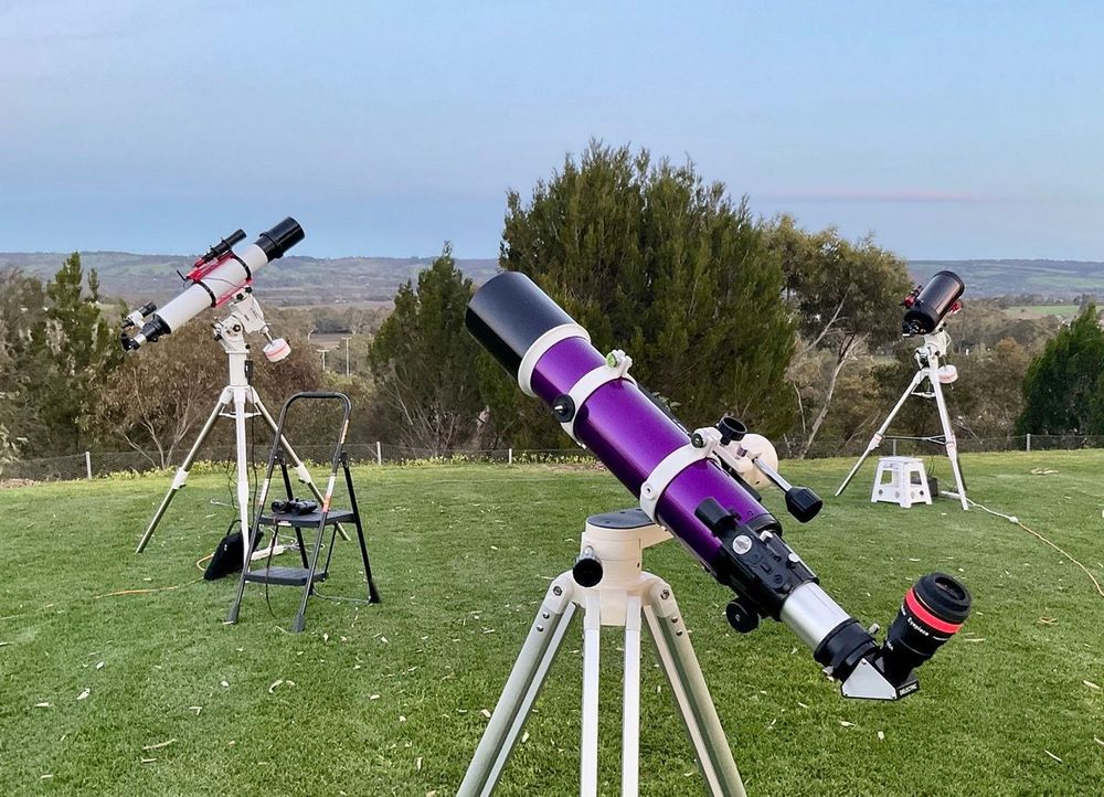 A photograph showing 3 telescopes set up for visual astronomy on a large lawned area with some trees and a hillside in the distance.