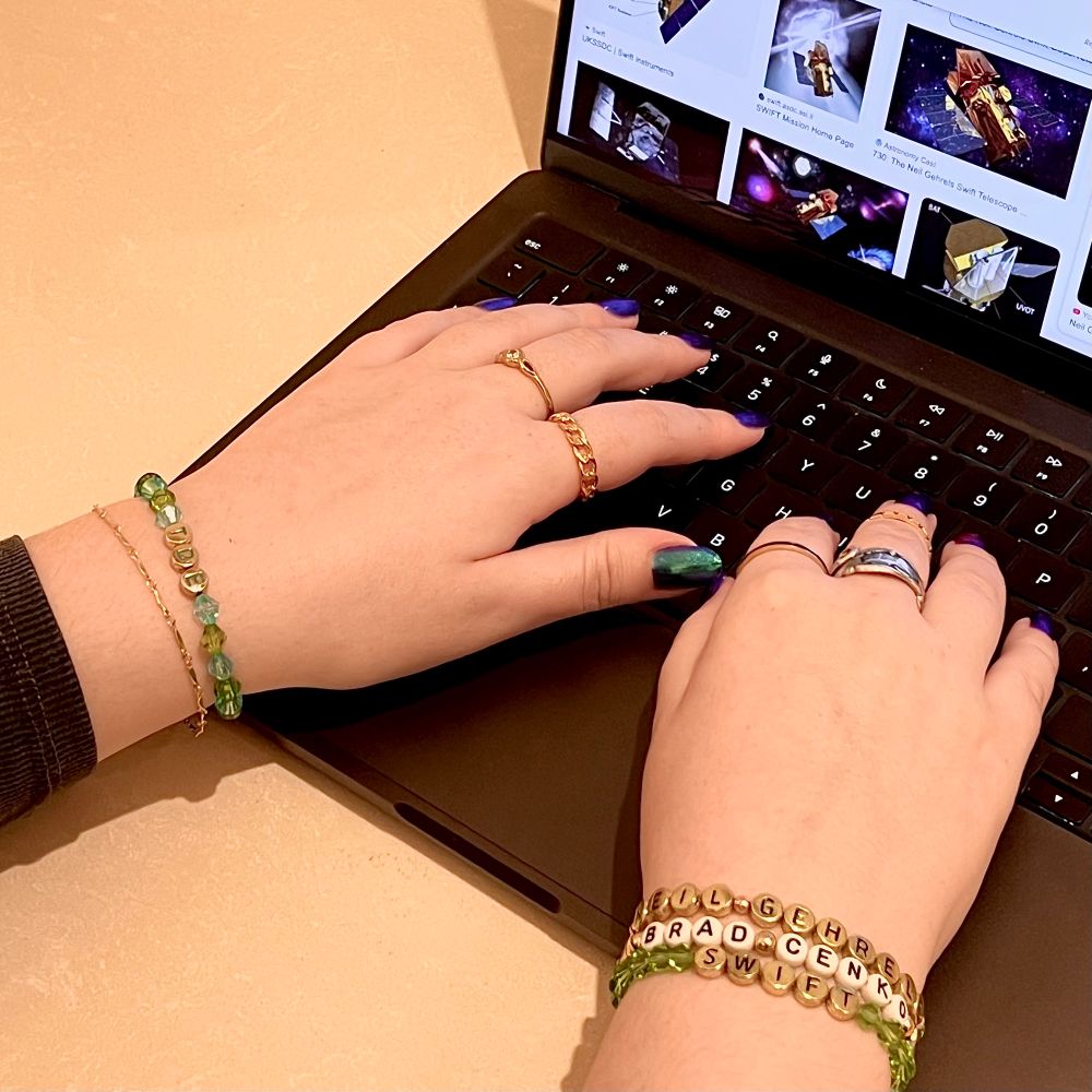 Two hands typing on a laptop.  On the screen is a image search engine result showing the Swift Observatory.  On the hands are 4 bracelets which say “TDE,” “Neil Gehrels,” “Brad Cenko,” and “Swift.”