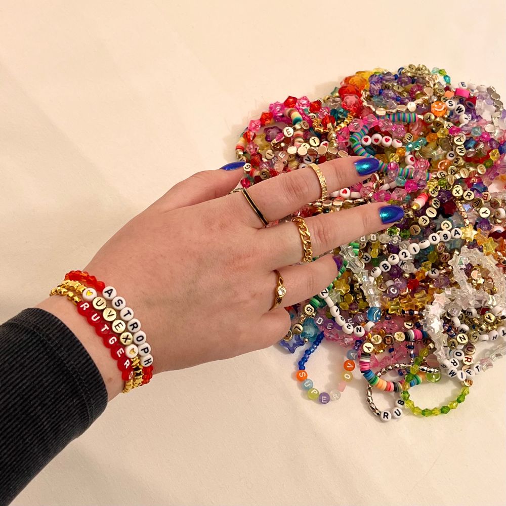 A hand reaching toward a pile of colorful friendship bracelets.  On the wrist of the hand there are three red, white, and gold bracelets that say “Astroph,” “UVEX,” and “Rutgers.”