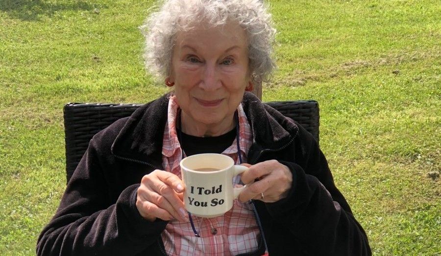 Margaret Atwood sitting outside in a garden chair with green grass in the background. She's holding a cup that reads "I told you so". A beautiful knowing smile on her face