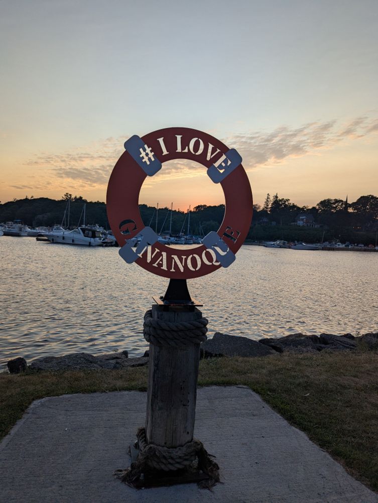 A metal sign in the shape of a life preserver against a sunset on the St Lawrence River. Inscribed in the sign is #I love Gananoque 