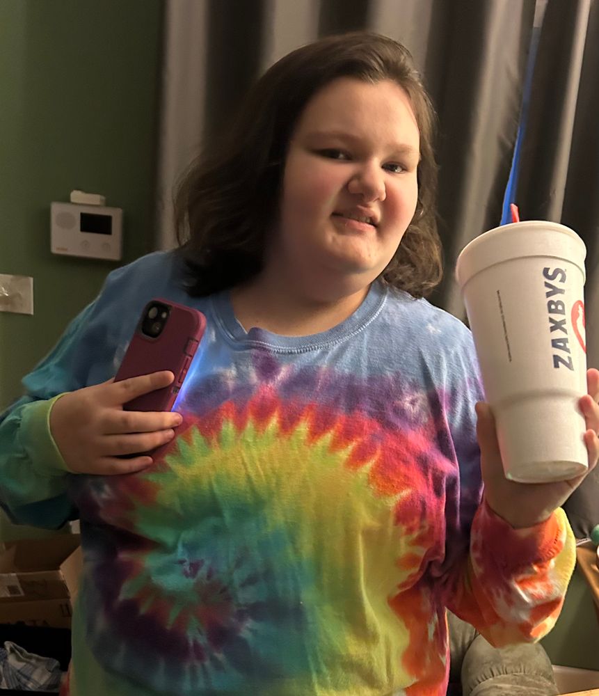 A teen girl wearing a tie dye long sleeve shirt, while holding a deep purple iPhone and a large Zaxby’s cup
