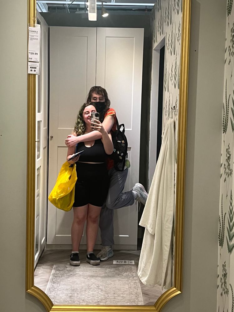 me (right) and my beautiful transsexual girlfriend (left) pose in a mirror at ikea. she has one leg kicked up and her arms around me. im holding her phone and a big yellow bag full of doodads and plushies. she is carrying my bag for me. we are in love btw. 