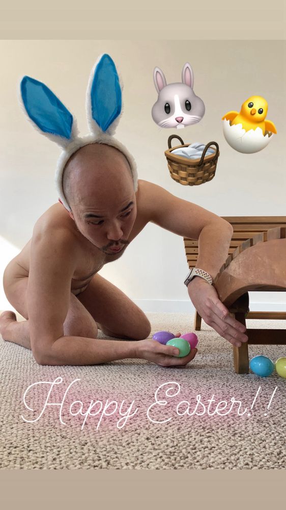 In naked man in bunny ears looking under chair and placing coloured eggs