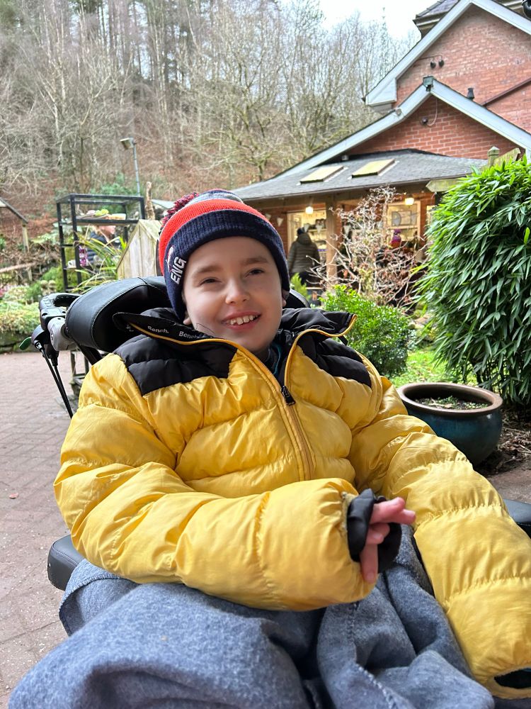 A smiling boy in a wooly hat and yellow winter coat sitting in an outdoor cafe in the woods  in a wheelchair 