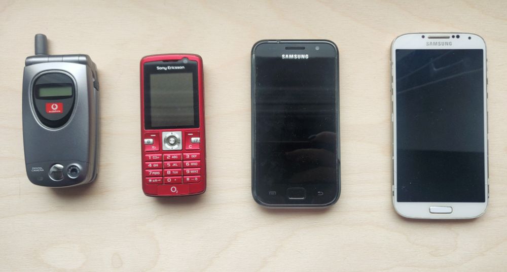 Vier Handys unterschiedlicher Bauarten auf einer hellen Fläche liegend. Von links nach rechts immer moderner und größer werdend. Von links nach rechts: ein silbernes Klapphandy von Sharp, ein rotes Handy von Ericsson, ein schwarzes Smartphone von Samsung, ein weißes Smartphone von Samsung. 