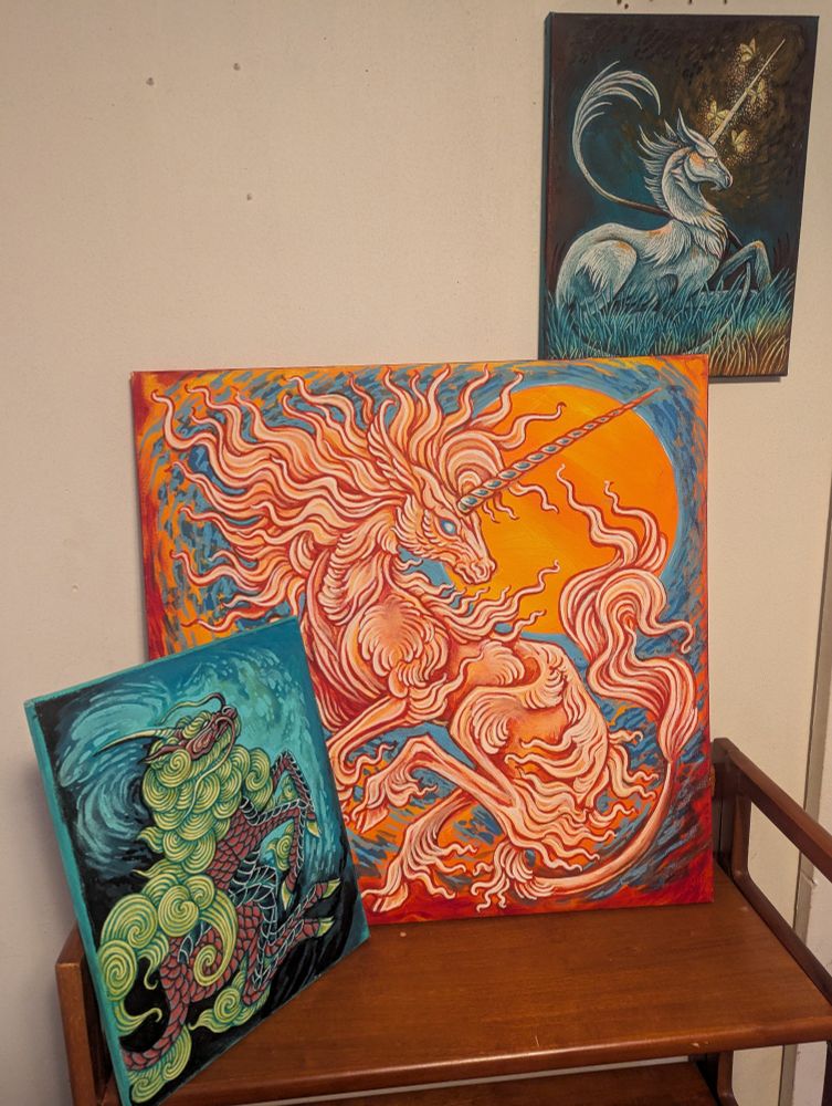 Three paintings on a bookshelf and a wall: one of a fiery unicorn, one of a green and bronze Kirin and one of a unicorn reclining with glowing butterflies around its horn. Go visit Adrian's Anomalies on Etsy, this is their work it's amazing.