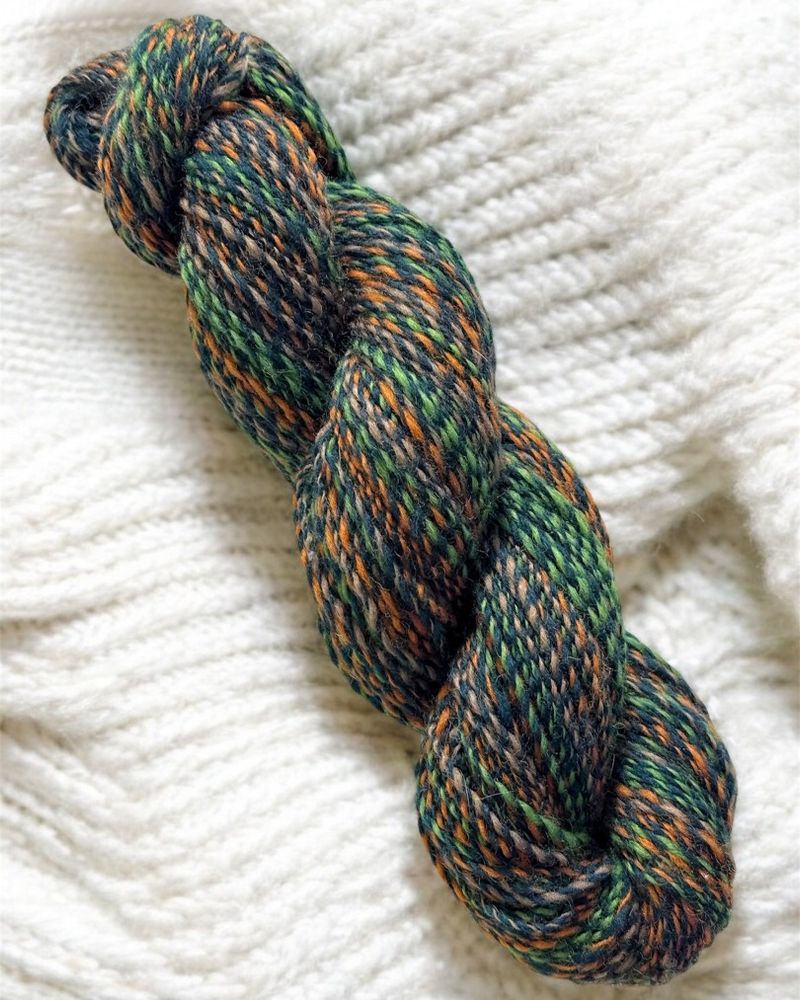 A handspun yarn skein featuring earthy tones of green, brown, and gold, reminiscent of a forest floor. The skein is coiled neatly against a soft white fabric background.