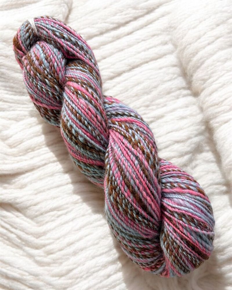 A close-up of a handspun yarn skein in the colorway 'Chocolate, Please,' featuring earthy brown tones interwoven with pink and blue accents, lying on a soft, creamy white background.
