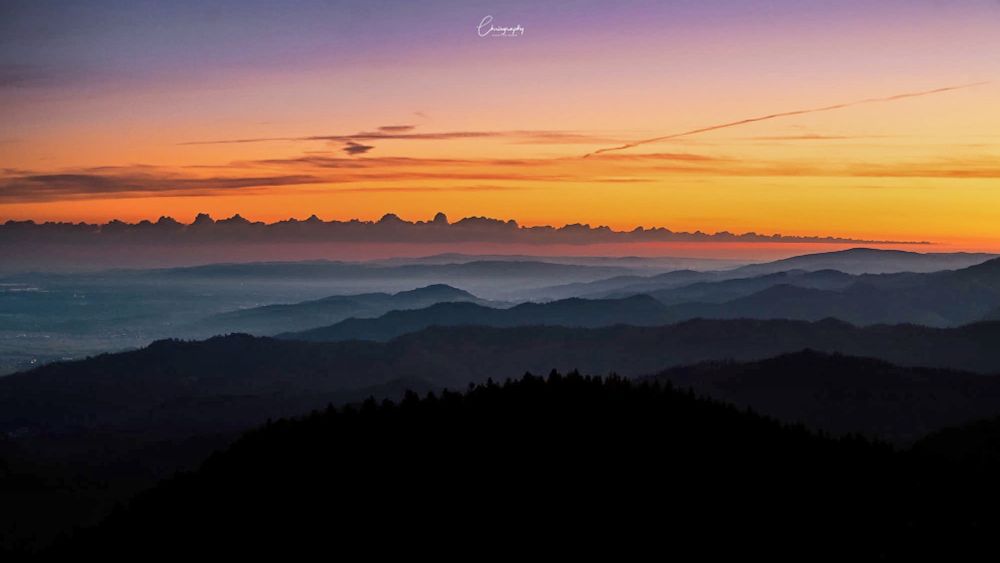 Sonnenaufgang über einer Berglandschaft mit mehreren silhouettenhaften Bergketten und einem farbenfrohen Himmel in Orange-, Lila- und Blautönen.