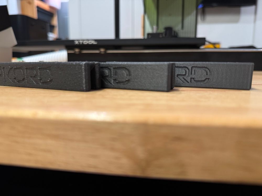 Three 3D-printed objects with embedded lettering sit on a workbench for comparison. From left to right, they were printed with a 0.8mm nozzle, 0.6mm, and 0.4mm. The leftmost looks rough and disfigured, the right looks nearly perfect.