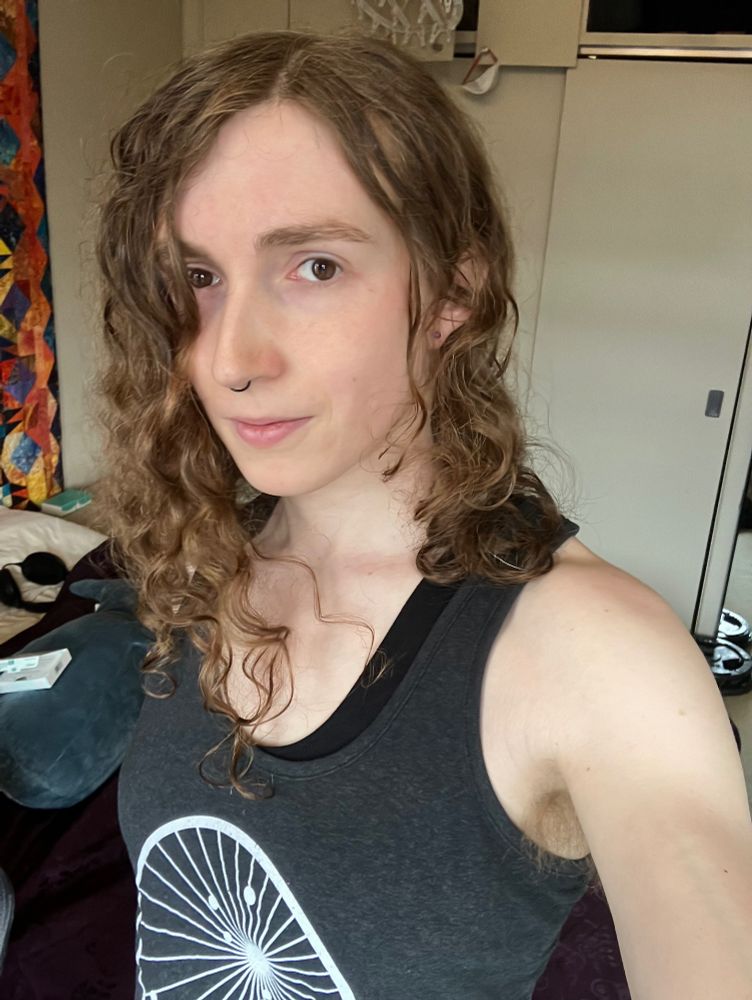 A selfie of a buff climber t girl (me!). She has curly medium brown hair that’s just past shoulder length. She’s wearing a tank top so the girlies can see her guns. In the background you can see oestrogen patches sitting on a blåhaj and weight plates