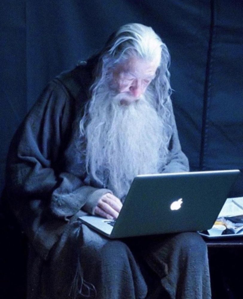 BTS photo from the filming of Lord Of The Rings. Sir Ian McKellen is in full costume and makeup as Gandalf, looking old AF, and is hunched over his macbook. He looks super old, tired, and a little sad