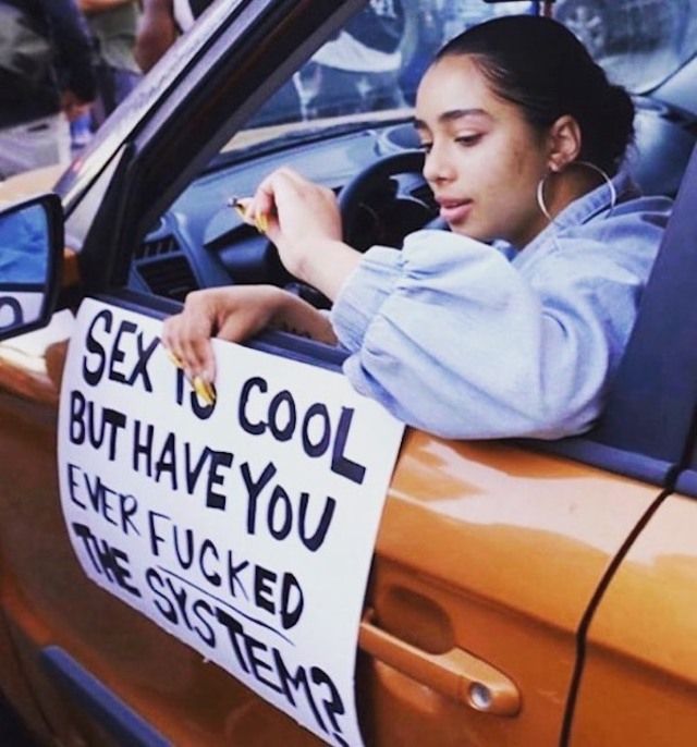 Junge Frau sitzt im Auto am Steuer, Fenster ist offen und sie hält ein selbstgemaltes Schild aus dem Fenster mit der Aufschrift "Sex is cool but have you ever fucked the system?"