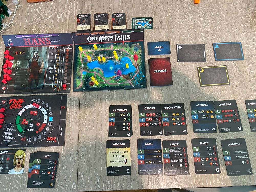 A table set up with the board game “Final Girl”. The Camp Happy Trails map, Hans killer board, and Laurie card are on the right. 11 action cards are spread out on the right.