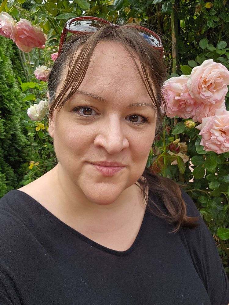 A woman with brown hair and brown eyes looks at the camera. Her sunglasses are on top of her head and she's squinting a little because of the bright light. She is standing in front of pale pink climbing roses.