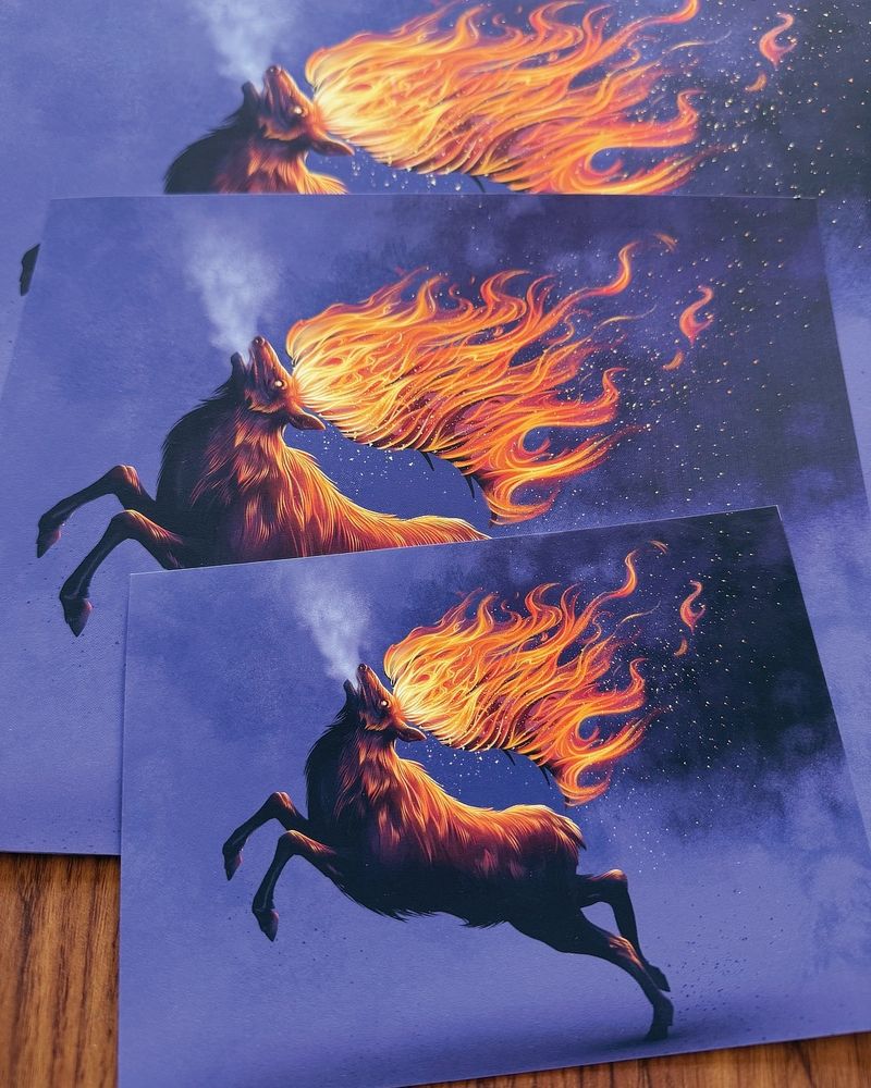 This is a photograph of several prints on a table. They depict an elk, running with its antlers on fire, over a purple background.