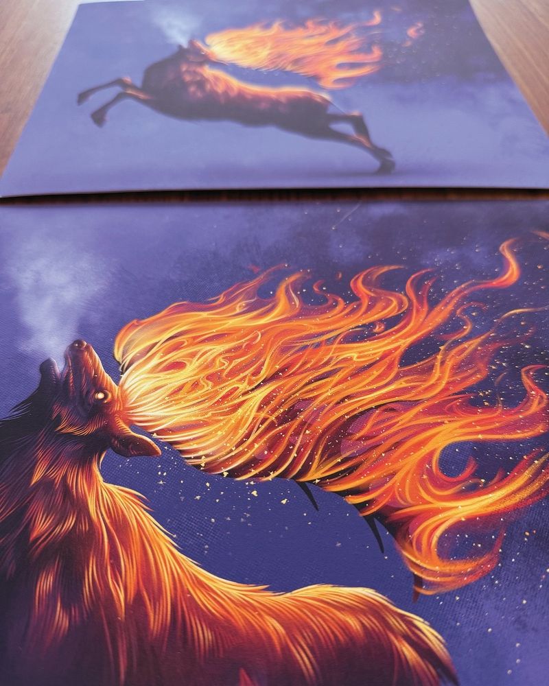 This is a photograph of several prints on a table. They depict an elk, running with its antlers on fire, over a purple background.