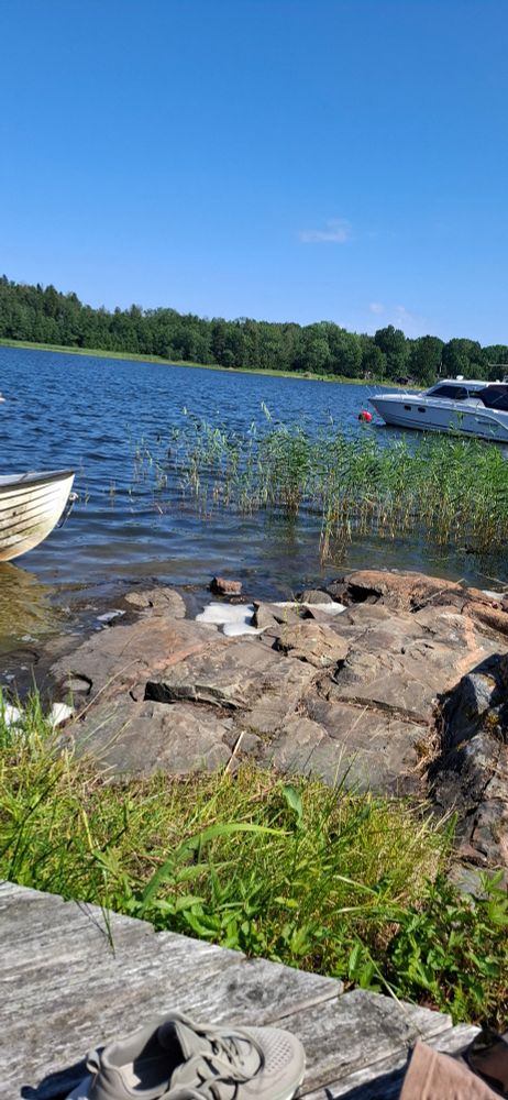 View of a small cove in sweden on the Baltic sea. There is a jetty with a rowboat visible, and another boat as well as granite rocks and reeds. In the background the forest is visible behind the water. A beautiful blue sky can also be seen