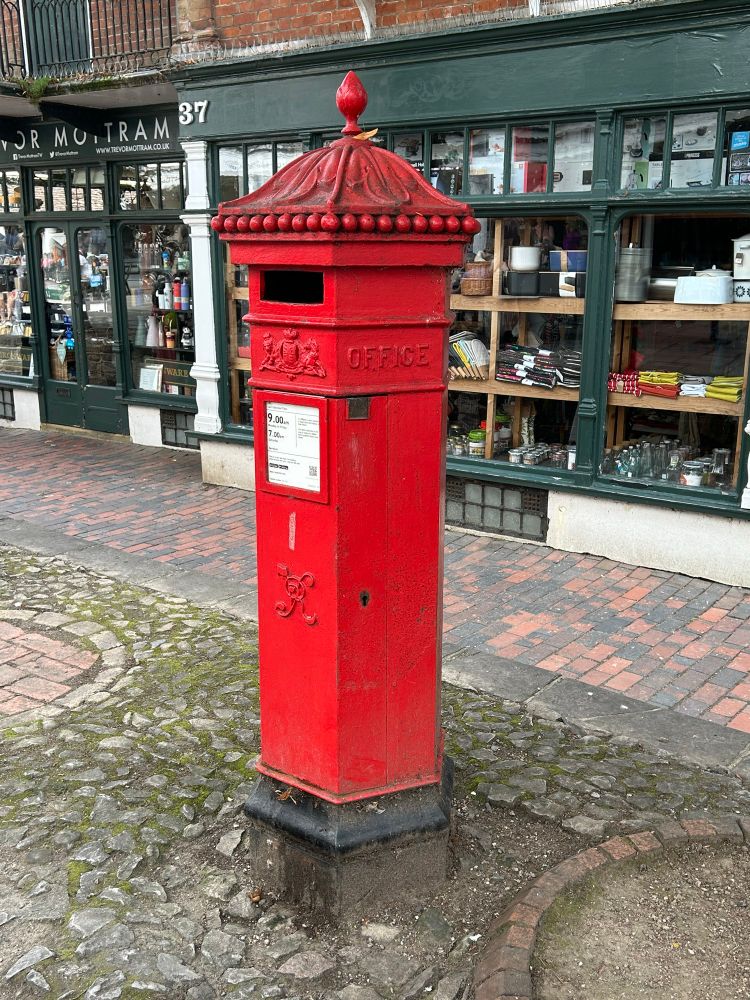A hexagonal red post box located on a cobbled pavement. A cookware shop is in the background. 