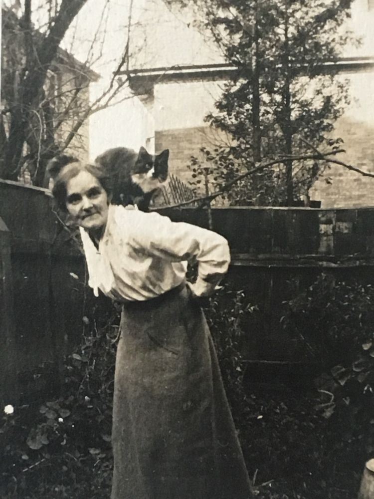 An old photo of a woman bending forward with a black and white cat sitting on her shoulder. 