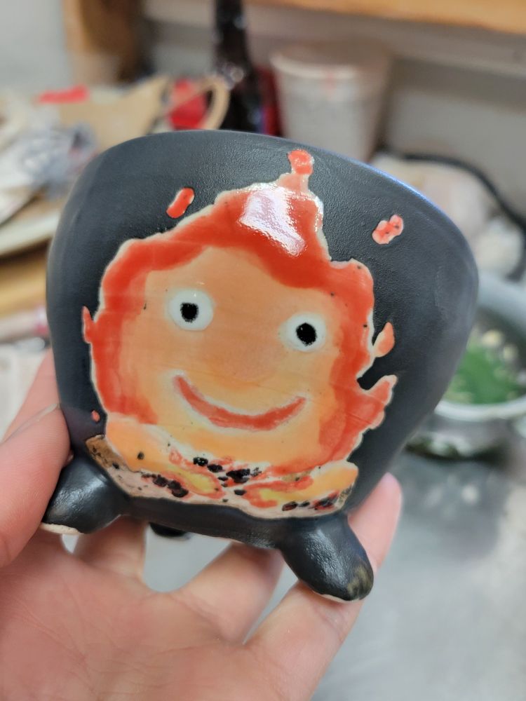 A black ceramic bowl with three small feet. The front is decorated with an image of Calcifer from "Howl's Moving Castle".