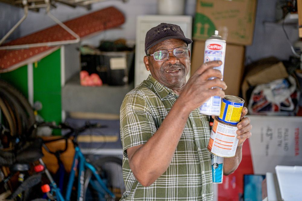 An older man wearing a plaid shirt and a cap holds several cans of spray paint in a garage filled with bicycles, boxes, and tools.
