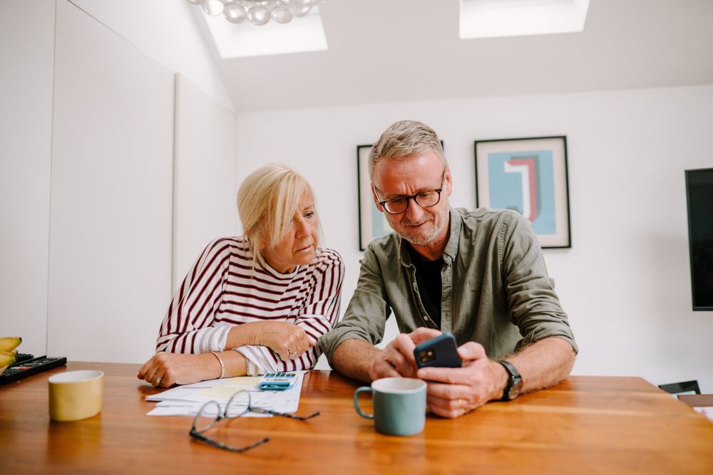 A older man and woman sit at a wooden table, reviewing bills and using a smartphone. Paperwork, glasses, and mugs are scattered on the table. The lighting is bright. The background shows framed artwork and a minimalist interior design.