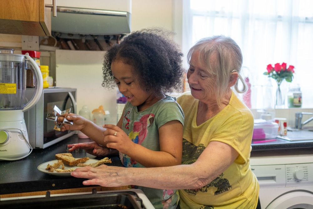 An older woman wearing a yellow tshirt makes toast with her granddaughter who is wearing a green tshirt. They are laughing in the kitchen as the cut the toast into star shaped slices.