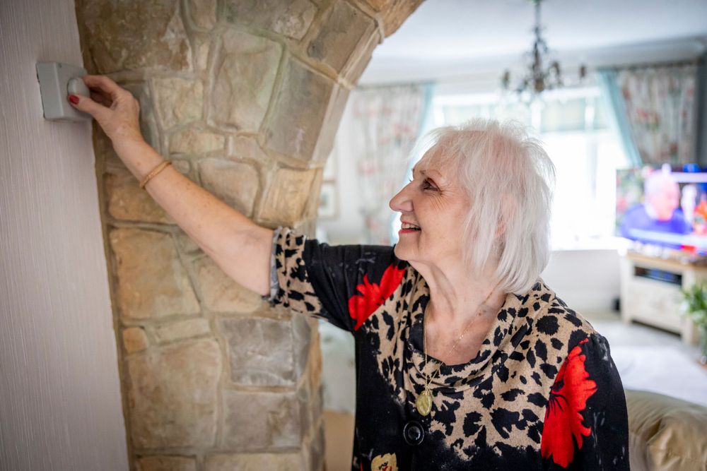 An older woman is seen in a bright living room, adjusting a thermostat on the wall. The thermostat is next to a stone archway. She is smiling, dressed in a leopard print blouse with red floral accents.