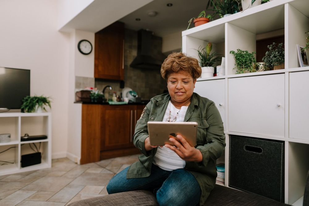 Older woman at home while using a tablet. She is sitting on a sofa in a living room with a modern interior design. The kitchen is in the background, featuring wooden cabinets. Shelves with storage bins and indoor plants decorate the room.  She is wearing jeans and a green shirt.