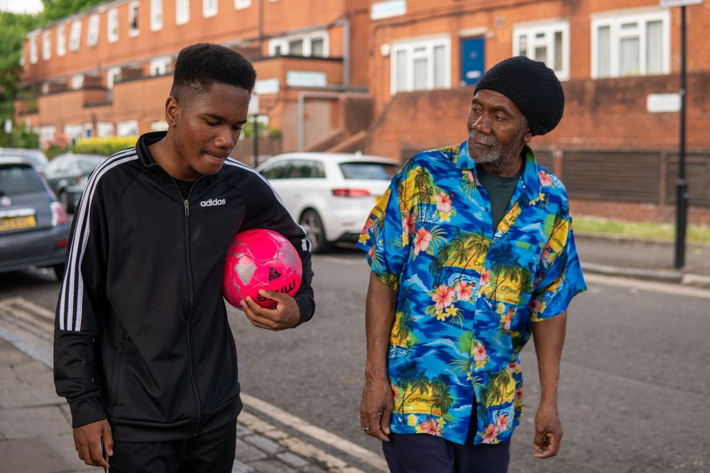 A father walks down the street with his son who is holding a pink football as the two converse.