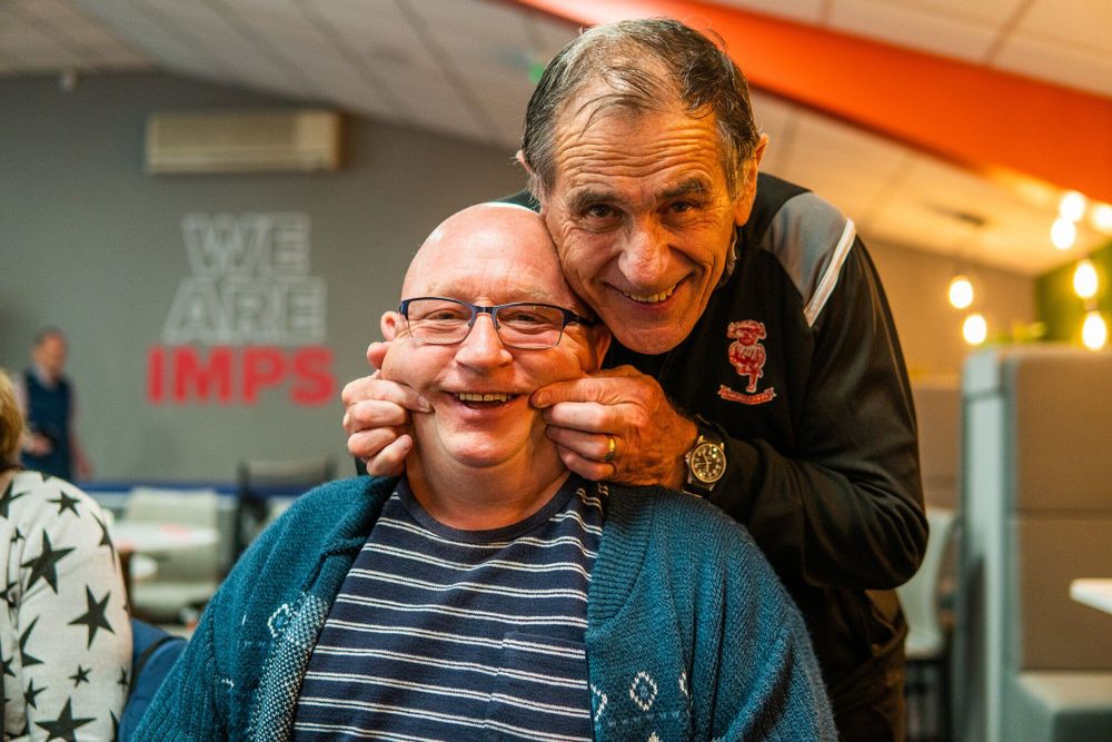 Two older men smile in a room, the man standing up is pulling the cheeks of the man sitting down into a smile. They are both happy.