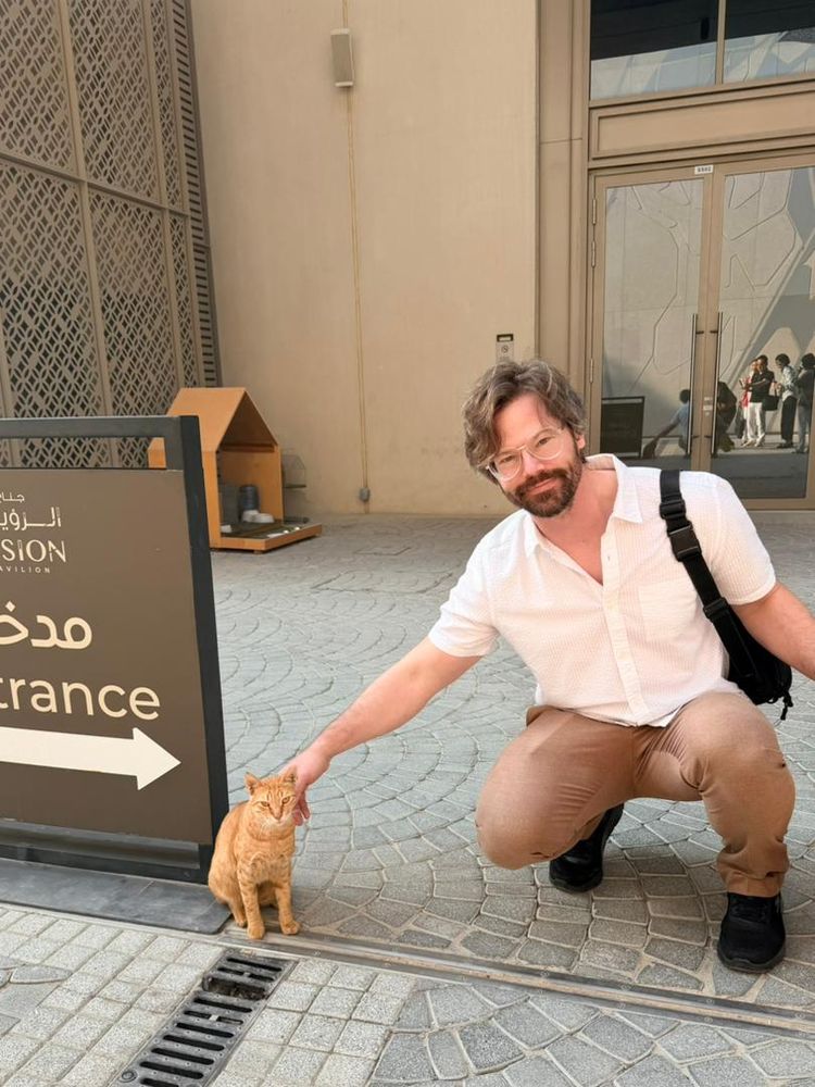 An orange cat appraises the camera with the calm confidence of place one sees in a lion. He is seated on a stone-paved area outside a building. Behind him a small pet house contains his food and water. A man cruoches next to him giving him a lil pet. A reflection over his shoulder shows the photographer and a retinue of onlookers