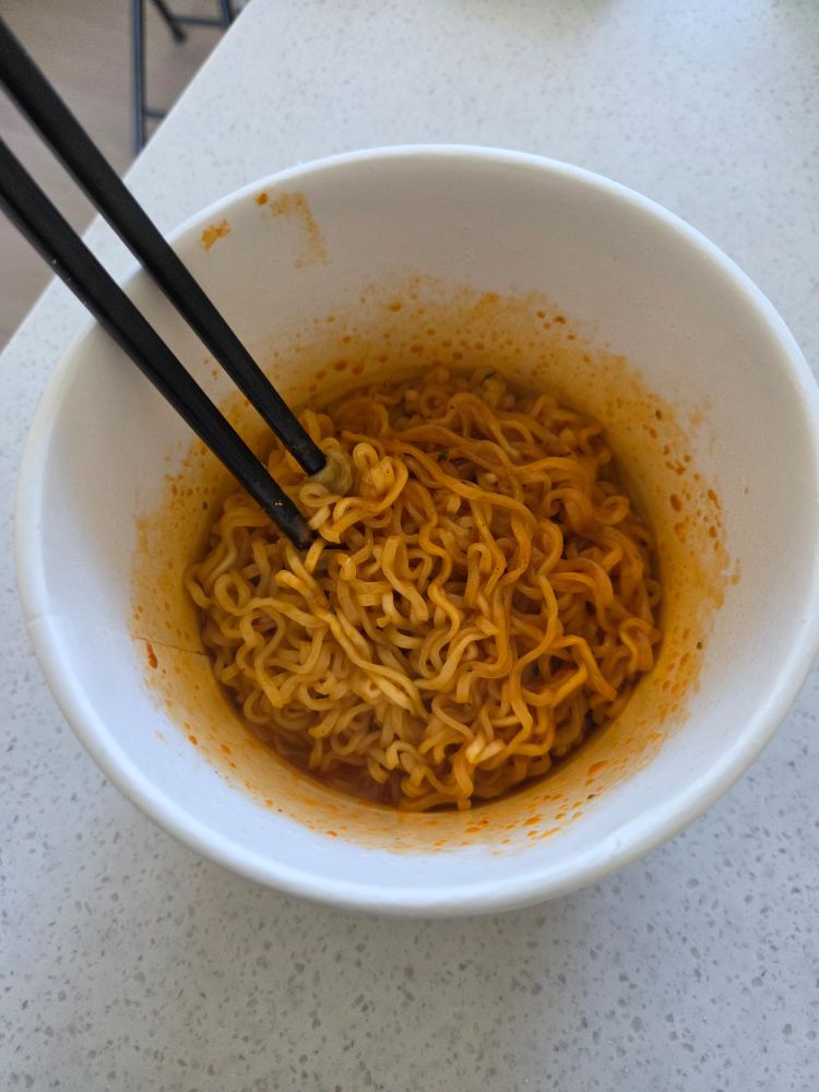 검은 나무 젓가락 한 벌이 꽂힌, 조리 이후의 푸팟퐁커리 불닭볶음면. 눈에 띄게 노란 양념에 비벼진 인스턴트 볶음면이 가득하다. 